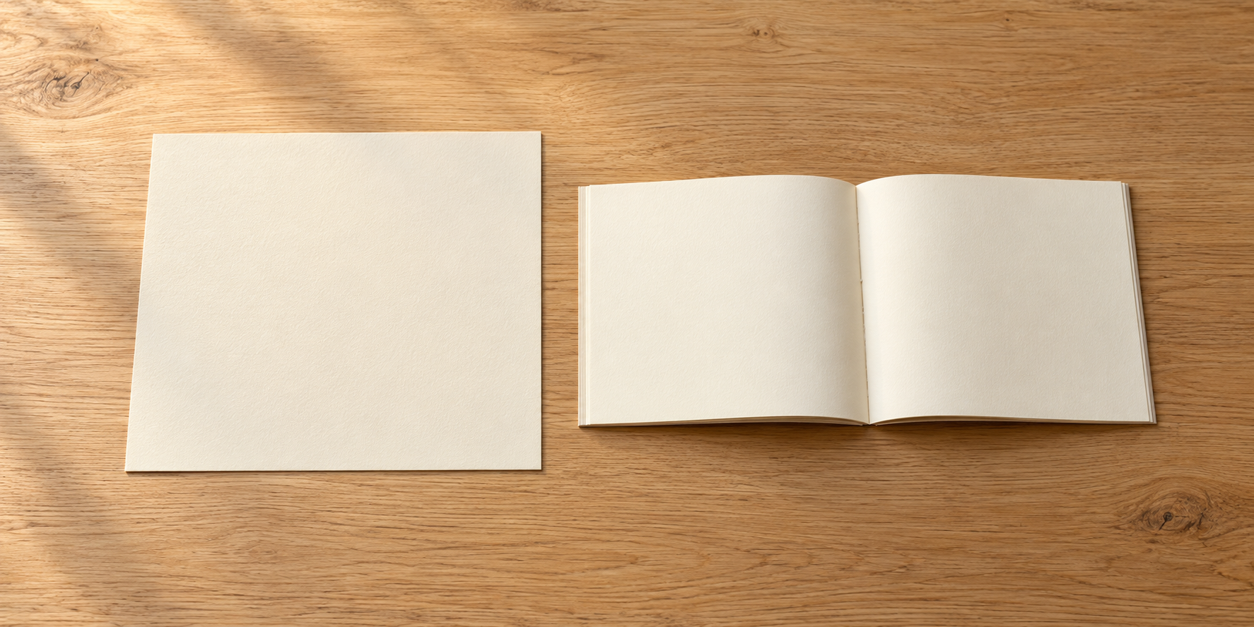 白い色紙と開いた無地の冊子を、木の机に離して並べている。
