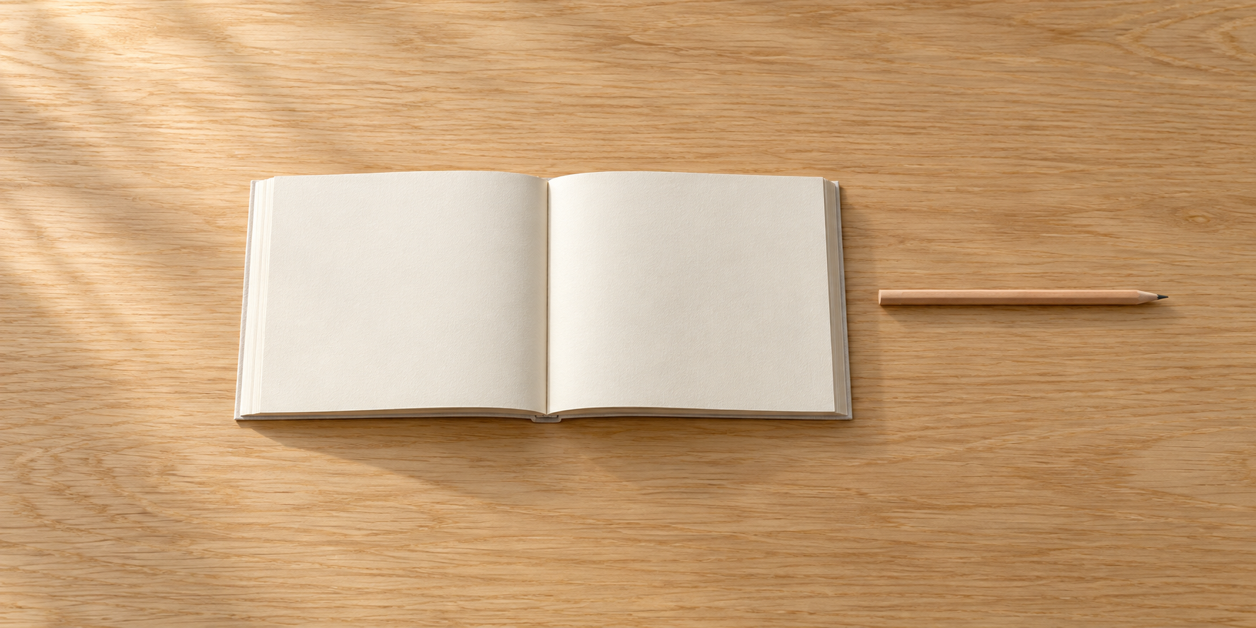 無地のページを開いた一冊の本と木軸の鉛筆を、木の机に置いている。