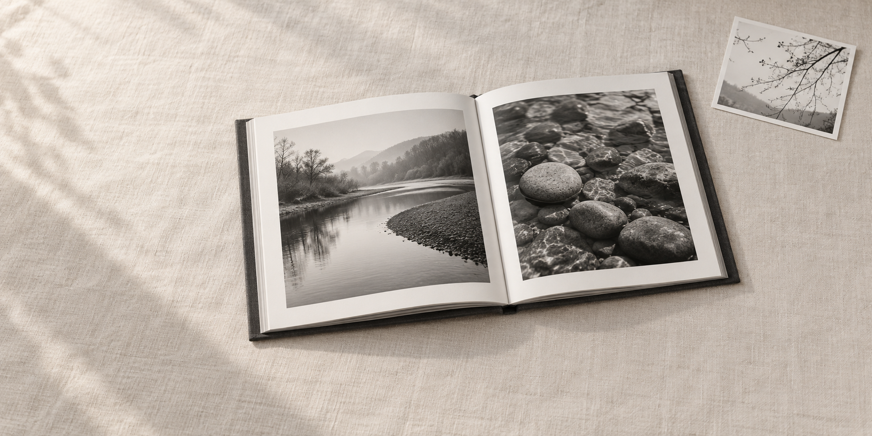 川岸と丸い石のモノクロ写真を載せた開いた本と、枝の小写真が布の上に置かれている